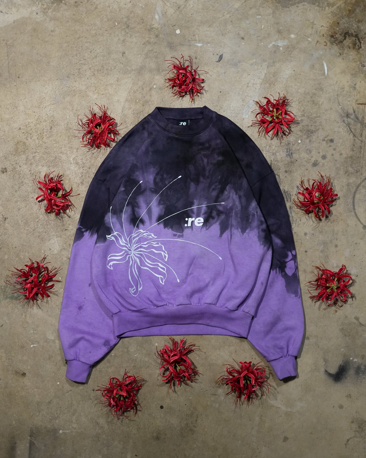 Purple and black tie-dye sweatshirt with a graphic design on a concrete surface with red flowers.